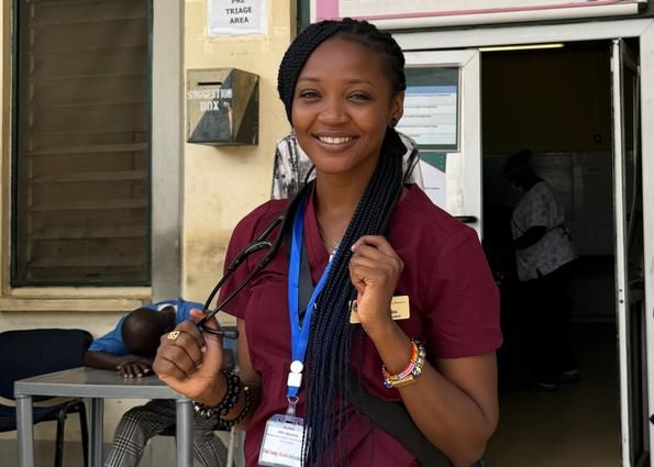 Hilda Agyapong, MPAS '24, wears maroon scrubs and a stethoscope standing outside a hospital entrance in Ghana.