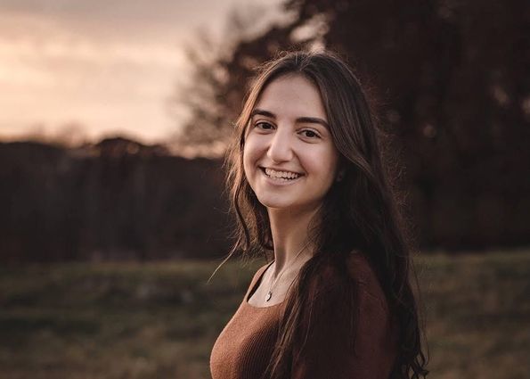 Ani Ourfalian, stands in a field and looks over her left shoulder to smile at the camera. She has long, dark brown hair and is wearing a lighter brown shirt.