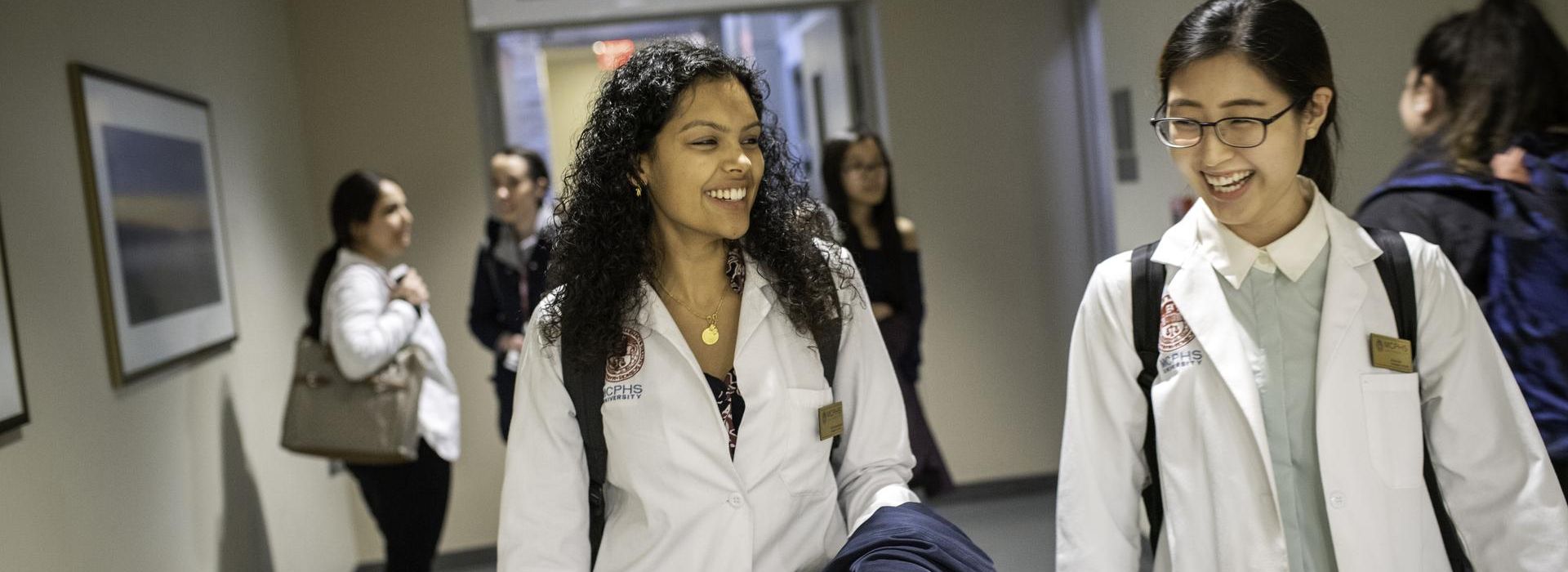 Two female students in white lab coats walking down hallway. 