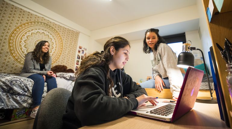 Three female students in a dorm room looking at a laptop. 