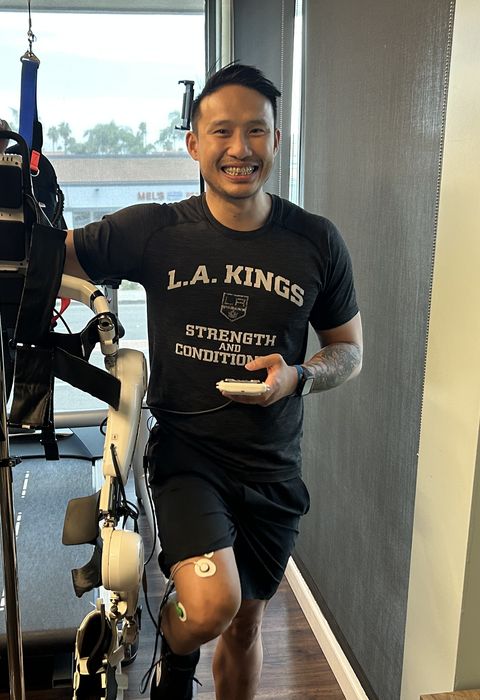 Michael Vu, DPT, smiles posing next to an assistive exoskeleton, a robotic device that connects via probes to patients' legs to help them walk.