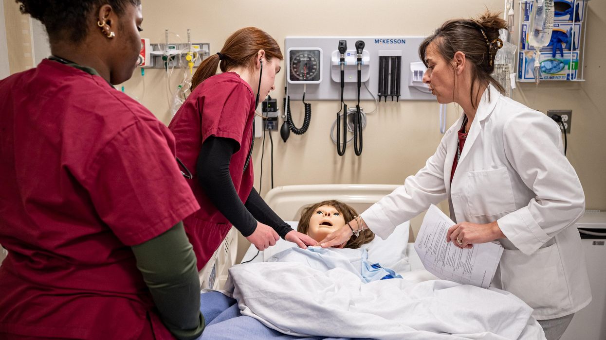 Nursing student in scrubs with stethoscope and teacher standing over mannequin in a bed.