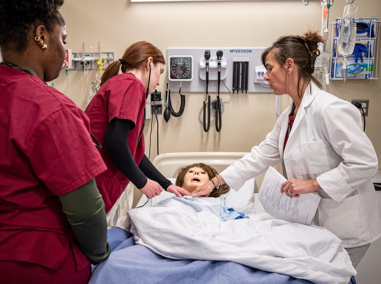 Nursing student in scrubs with stethoscope and teacher standing over mannequin in a bed.