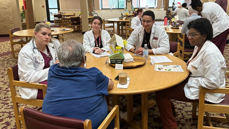 Healthcare students from MCPHS sit at a table talking with an older adult.