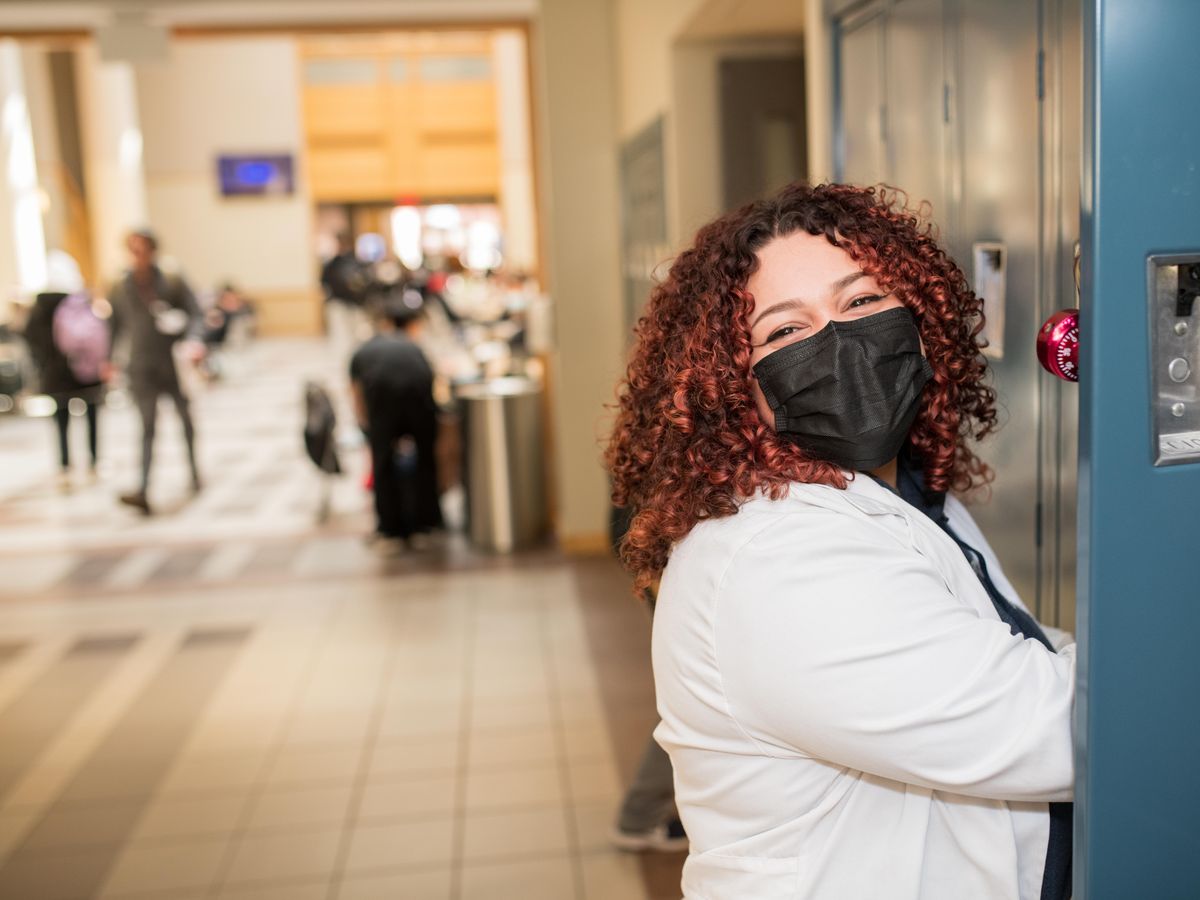 Smiling female student, wearing a mask in front of lockers. 