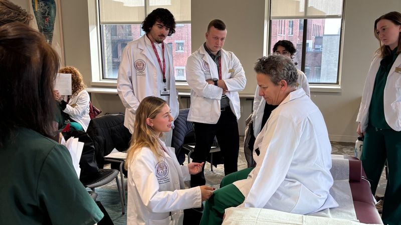 A physical therapy student from MCPHS demonstrates a treatment technique on an acupuncture student as a small group of others, all wearing white coats, listen,