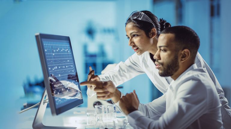 Man and woman in white coats pointing at a computer screen. 