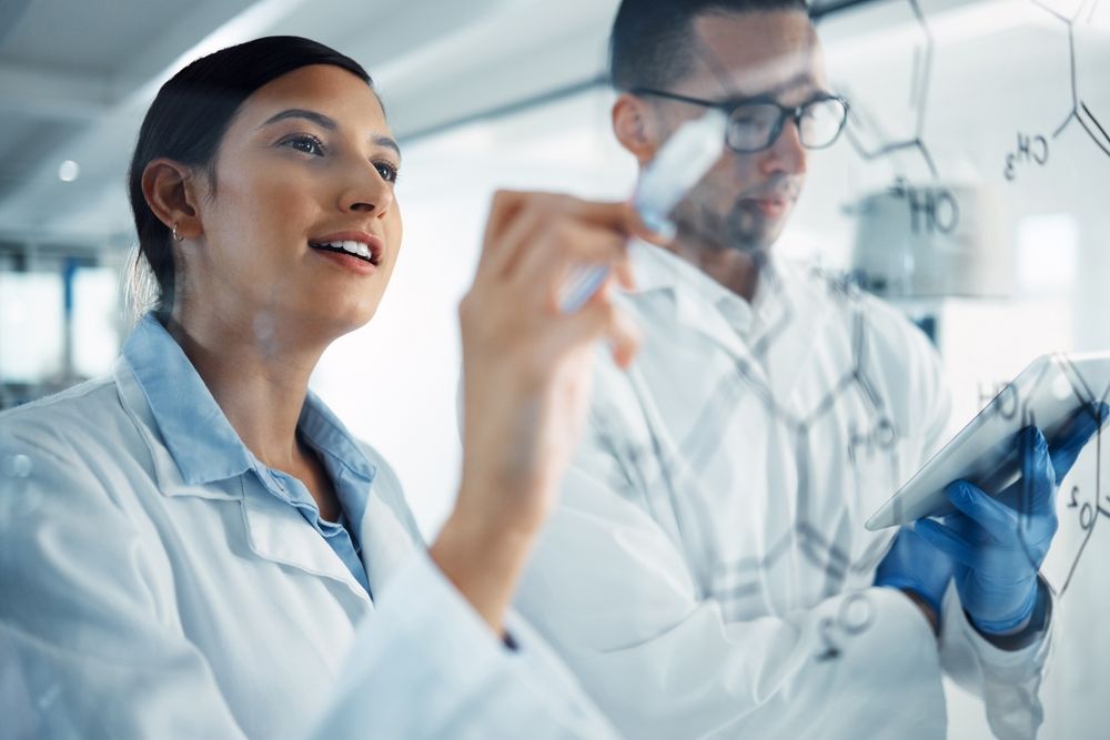 Two scientists in lab coats working at a glass board with chemical formulas written on it, while one holds a tablet.
