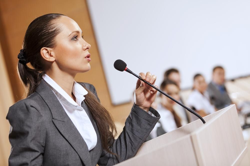 Woman in business attire at a podium, talking into a microphone. 