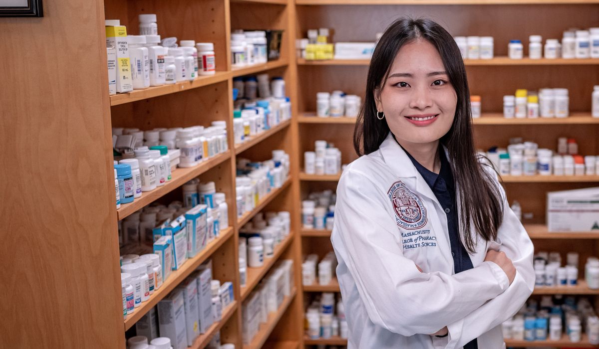 Smiling student in a white coat standing in a pharmacy lab. 