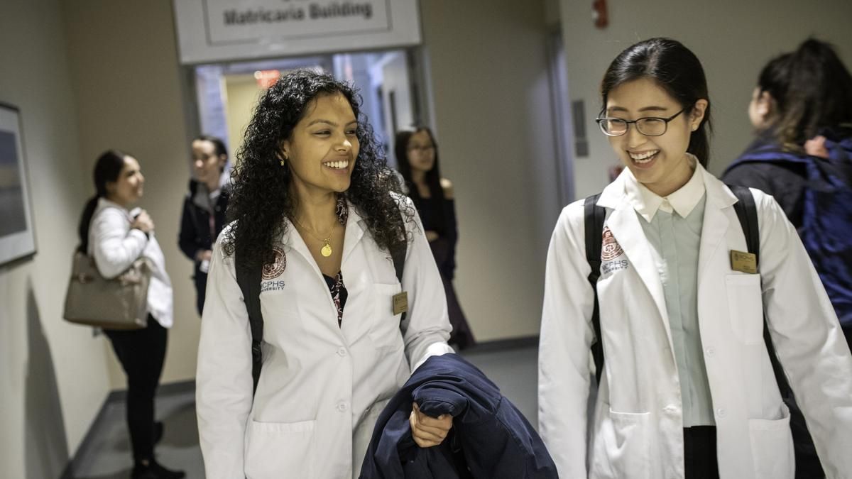 Two smiling female students in white coats walking down a hallway. 