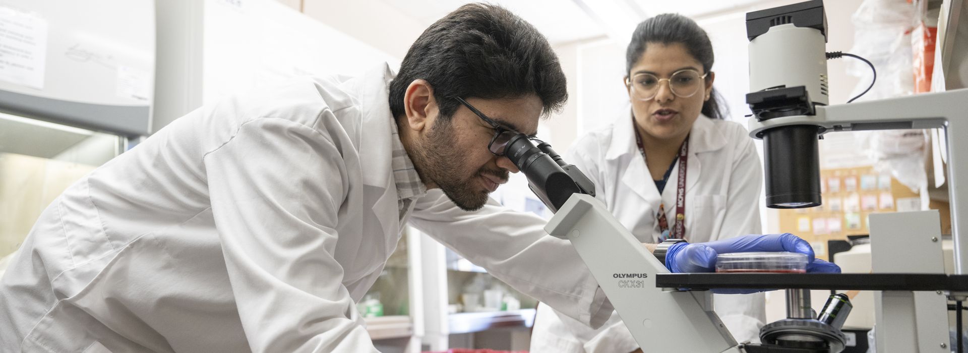 Two students in white coats, in a lab looking through a microscope