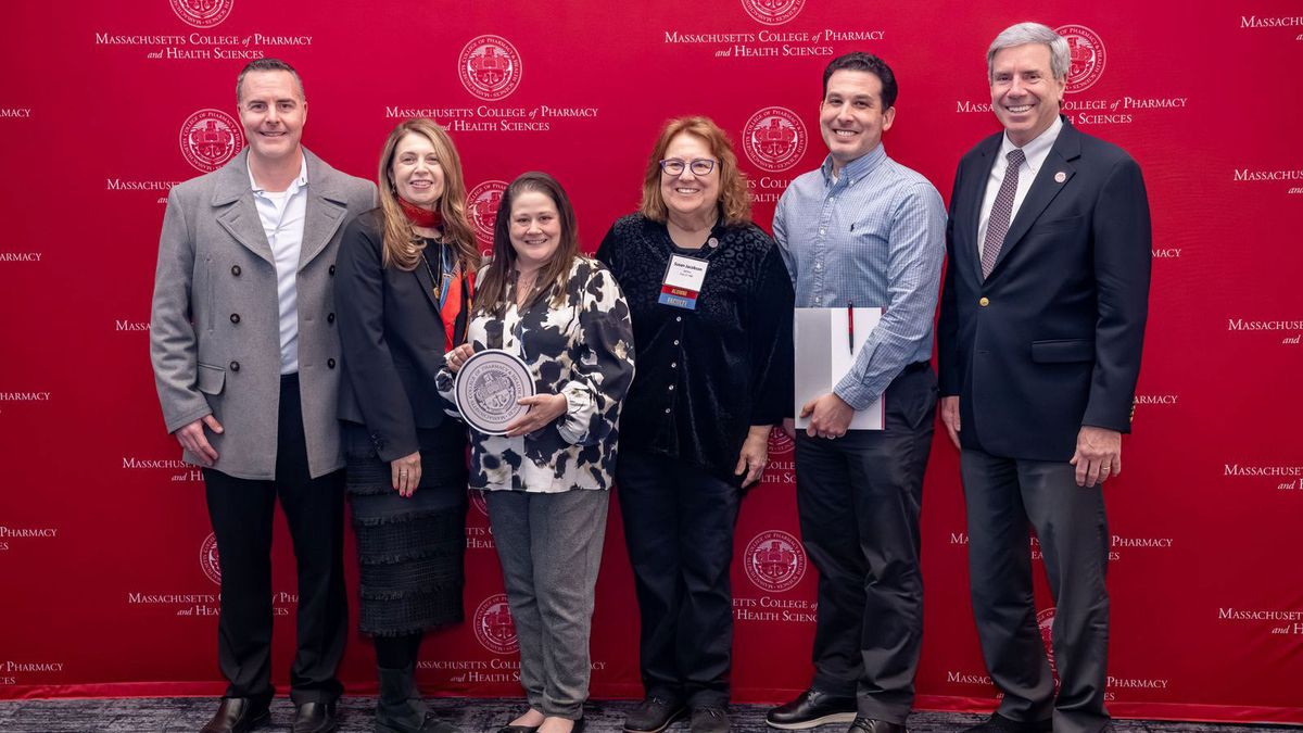 Faculty and Staff stand in front of an MCPHS red backdrop.