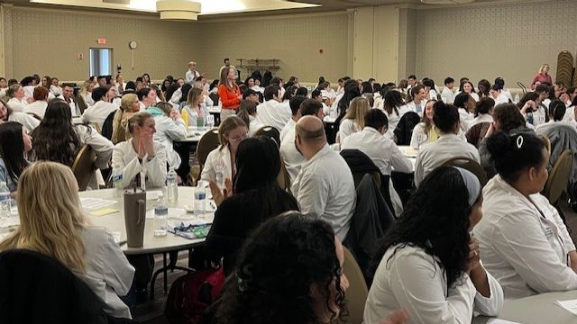 Healthcare students from MCPHS wearing white coats sit at round tables in a large conference room.