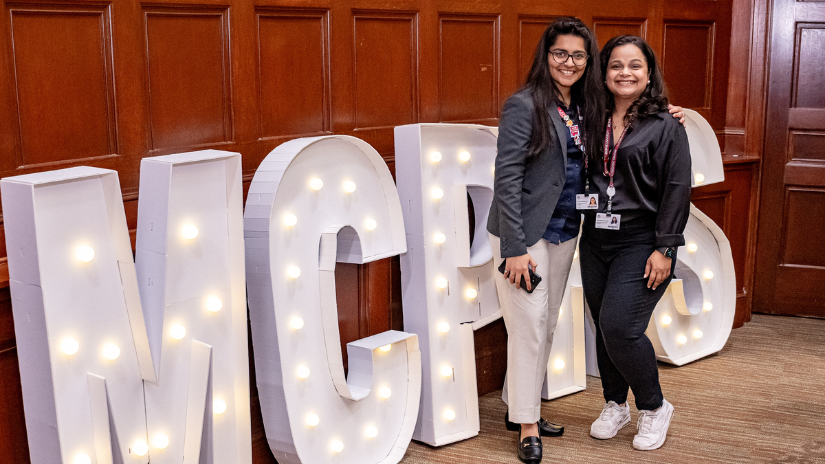 Two women stand in front of a MCPHS sign.