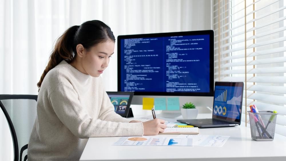 Female looking at a laptop and desktop with data on the screens. 