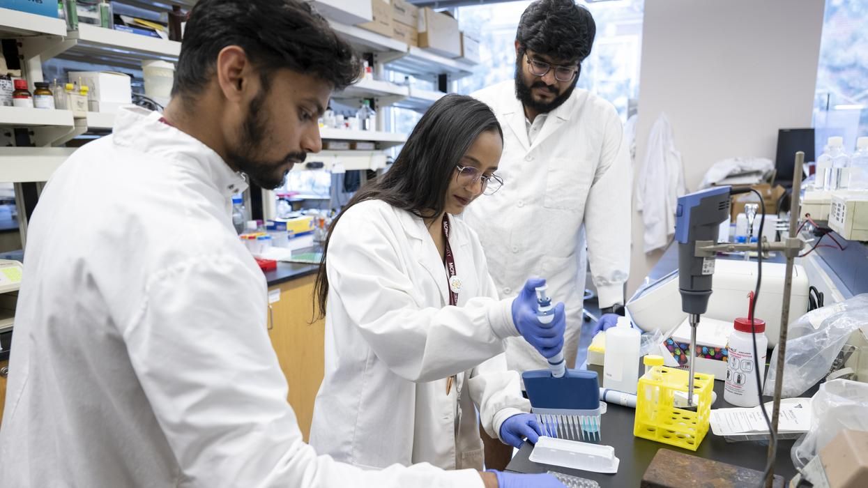 Three MCPHS pharmacy students in lab coats conducting experiments at a laboratory bench with scientific equipment and supplies.