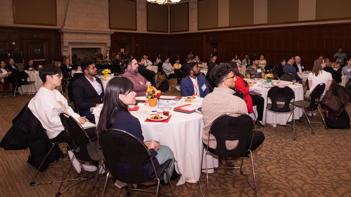 Students sit in a banquet hall at round tables with white tablecloths, turning their heads to listen to a presentation.