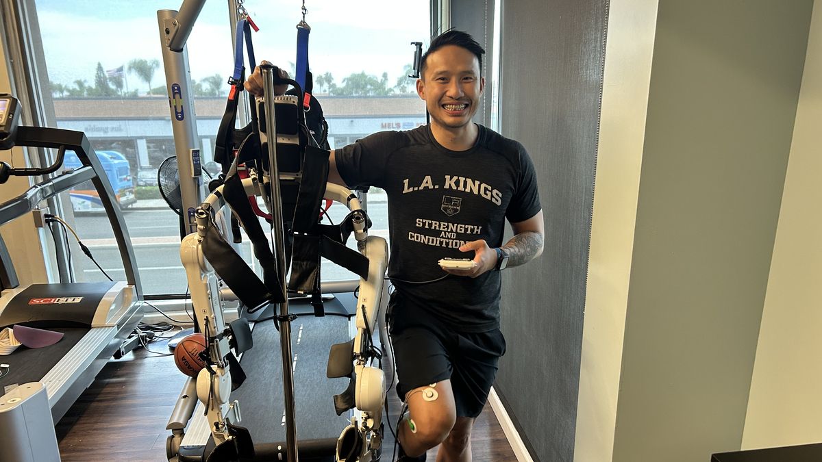 Michael Vu, DPT, smiles posing next to an assistive exoskeleton, a robotic device that connects via probes to patients' legs to help them walk.