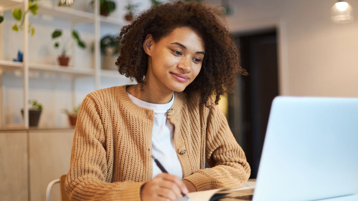 Female writing in a notebook with an open laptop in front of her. 