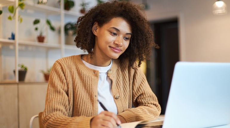 Female writing in a notebook with an open laptop in front of her. 