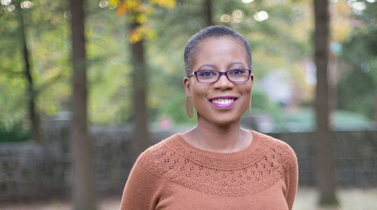 A close up of Nadia Obas who is smiling while wearing an orange sweater. She is standing in a forest with trees behind her that are out of focus.