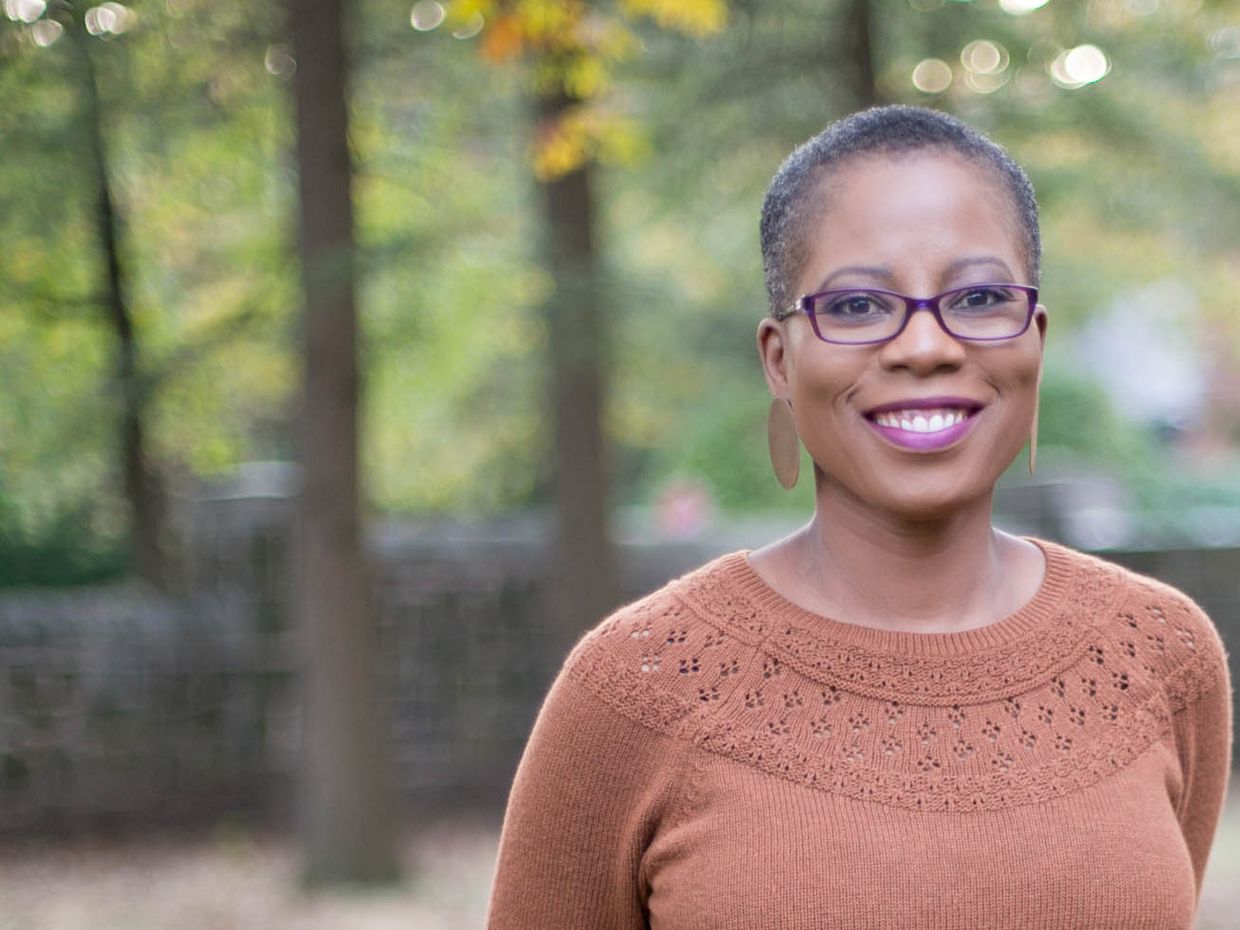 A close up of Nadia Obas who is smiling while wearing an orange sweater. She is standing in a forest with trees behind her that are out of focus.