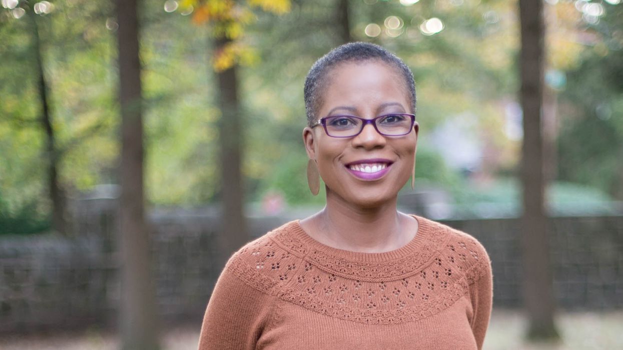 A close up of Nadia Obas who is smiling while wearing an orange sweater. She is standing in a forest with trees behind her that are out of focus.