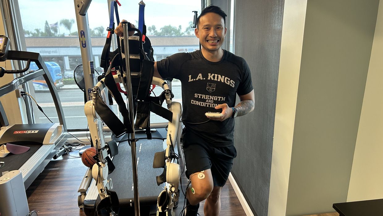 Michael Vu, DPT, smiles posing next to an assistive exoskeleton, a robotic device that connects via probes to patients' legs to help them walk.