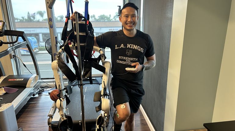 Michael Vu, DPT, smiles posing next to an assistive exoskeleton, a robotic device that connects via probes to patients' legs to help them walk.