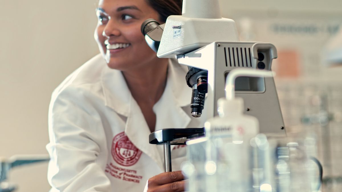 Smiling female student wearing a white coat in front of a microscope.