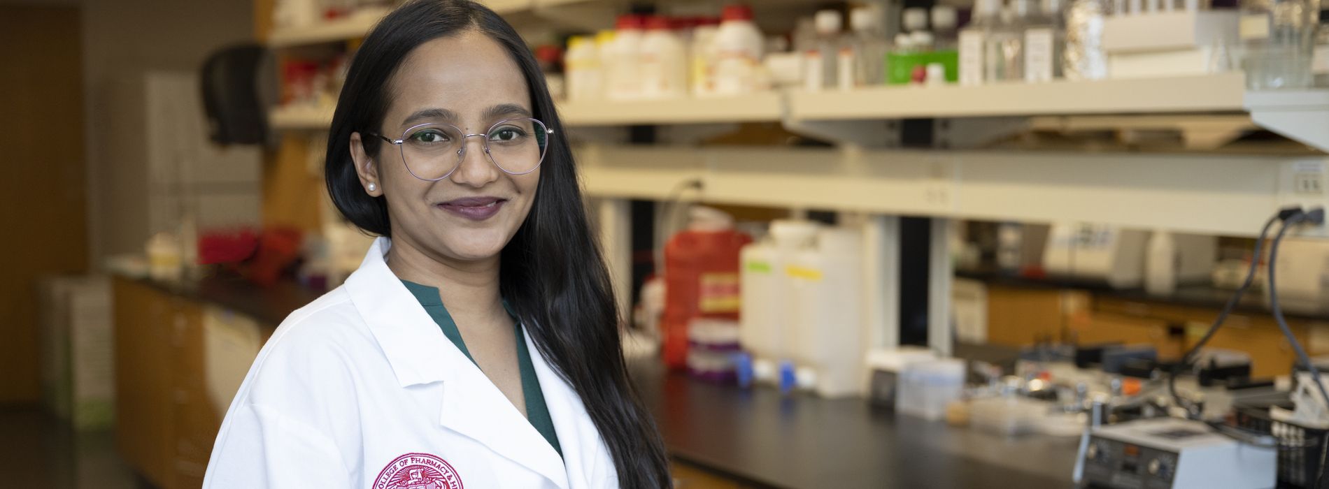 Smiling female student in white coat, standing in a pharmacy lab. 