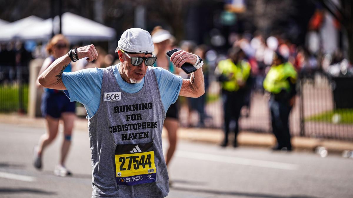 Bob Cargill, wearing sunglasses, a blue tank top with a grey t-shirt underneath, and a backwards white hat flexes both arms near his head while running the Boston Marathon.
