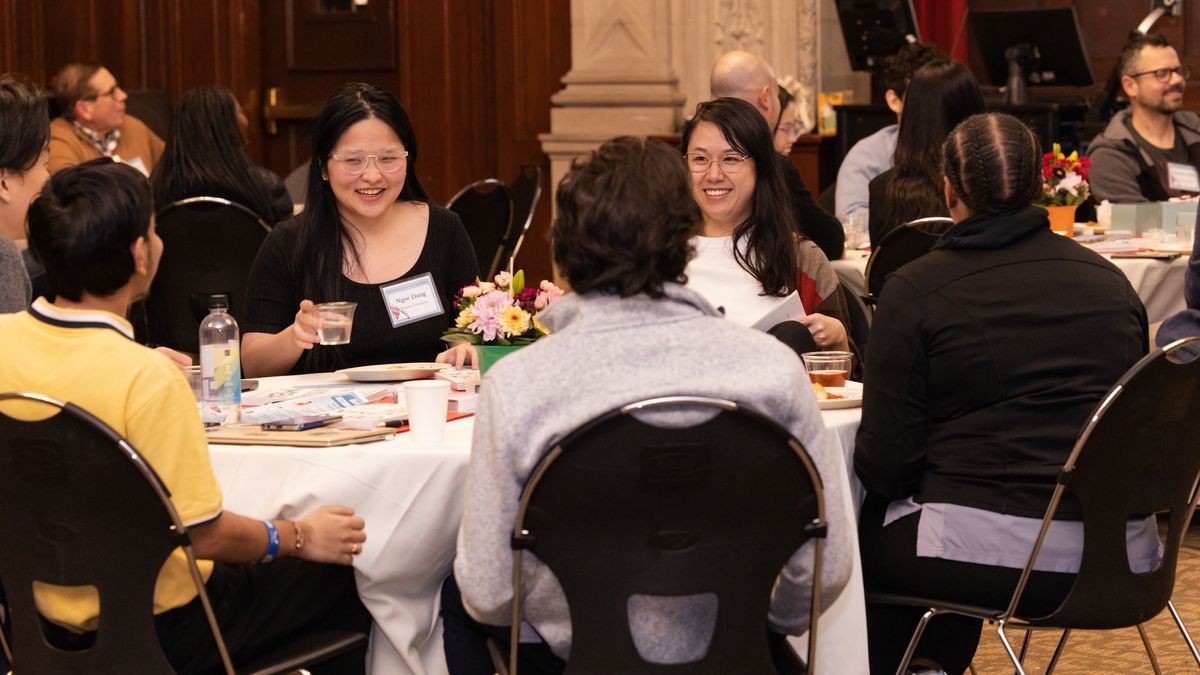A group of people sit at a round table, talking and interacting with each other.