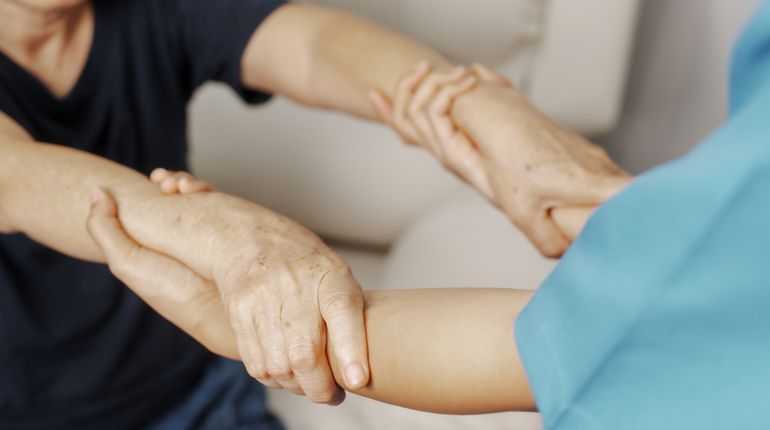 A healthcare provider holds the wrists of another person to help them stand up.