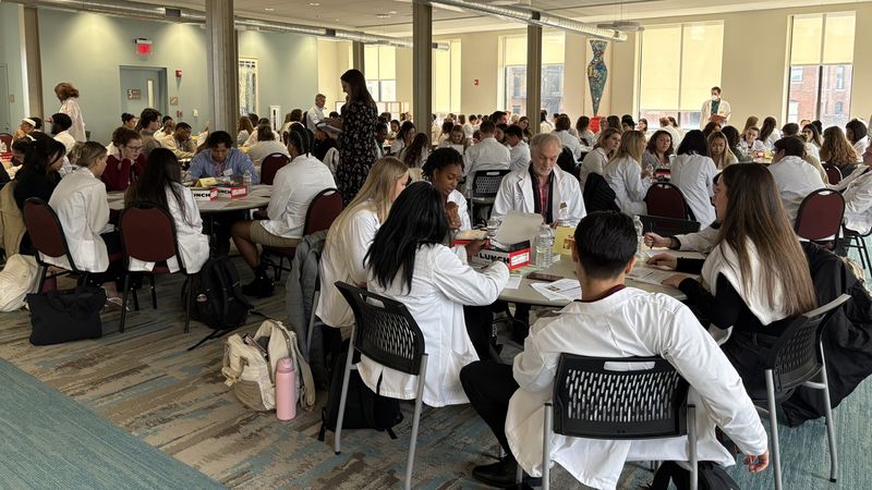 Healthcare students wearing white coats sit at round tables in a large conference room at MCPHS.