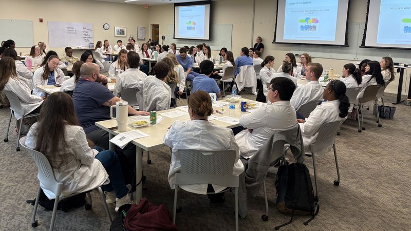 Healthcare students wearing white coats sit around tables talking in a large meeting room at MCPHS.