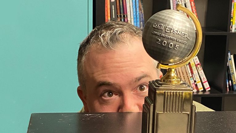 An image of A. David Lewis with only the top of his head and his eyes visible over a table as he peers at an award with a large globe on it. The globe has text that says "Will Eisner Comic Industry Awards 2005."