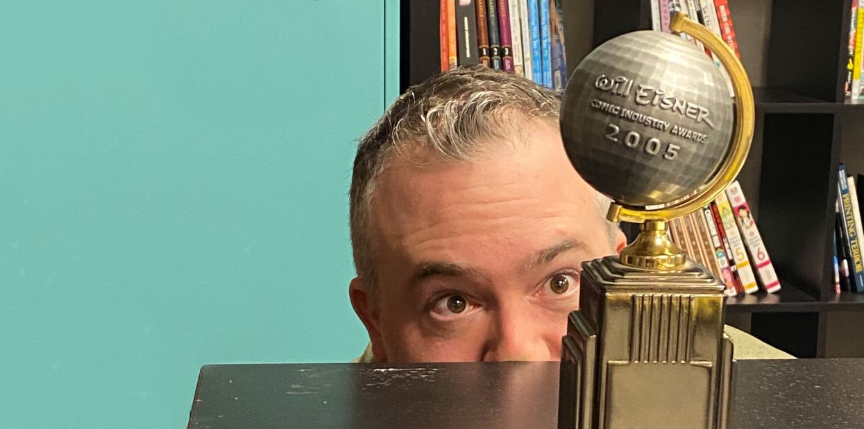 An image of A. David Lewis with only the top of his head and his eyes visible over a table as he peers at an award with a large globe on it. The globe has text that says "Will Eisner Comic Industry Awards 2005."