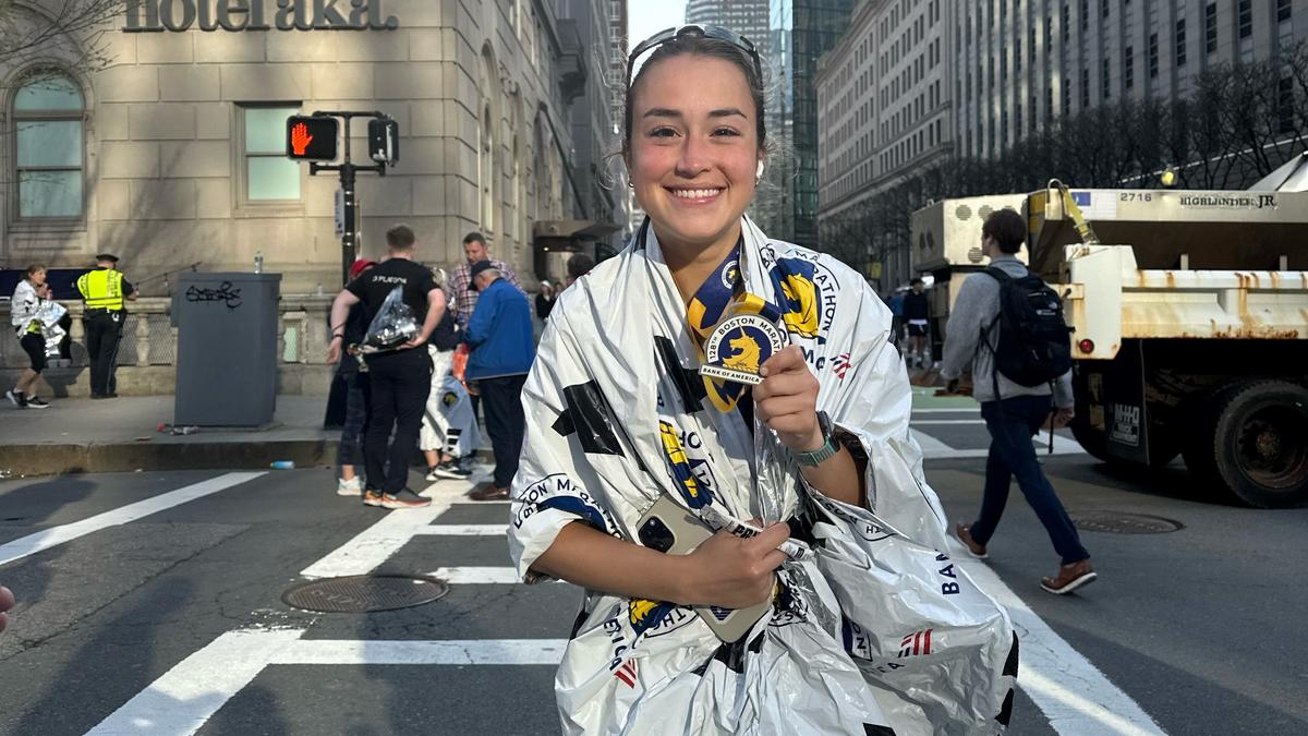 Holly Manzelli stands on a Boston street wrapped in a foil blanket while smiling and holding her Boston Marathon medal.