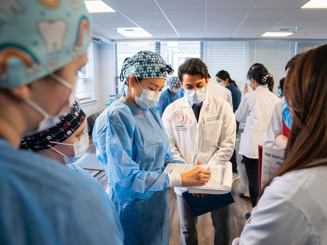 Students and staff in white coats and scrubs looking at a clipboard.